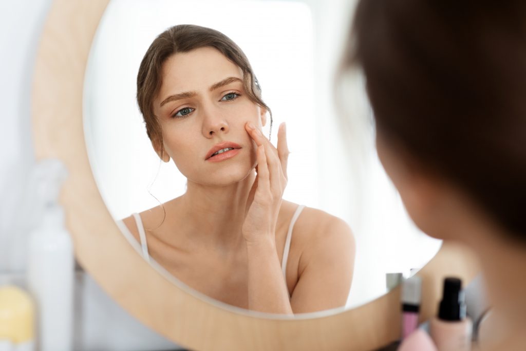 Acne prone skin concept. Upset young woman looking at mirror at home, checking her face skin