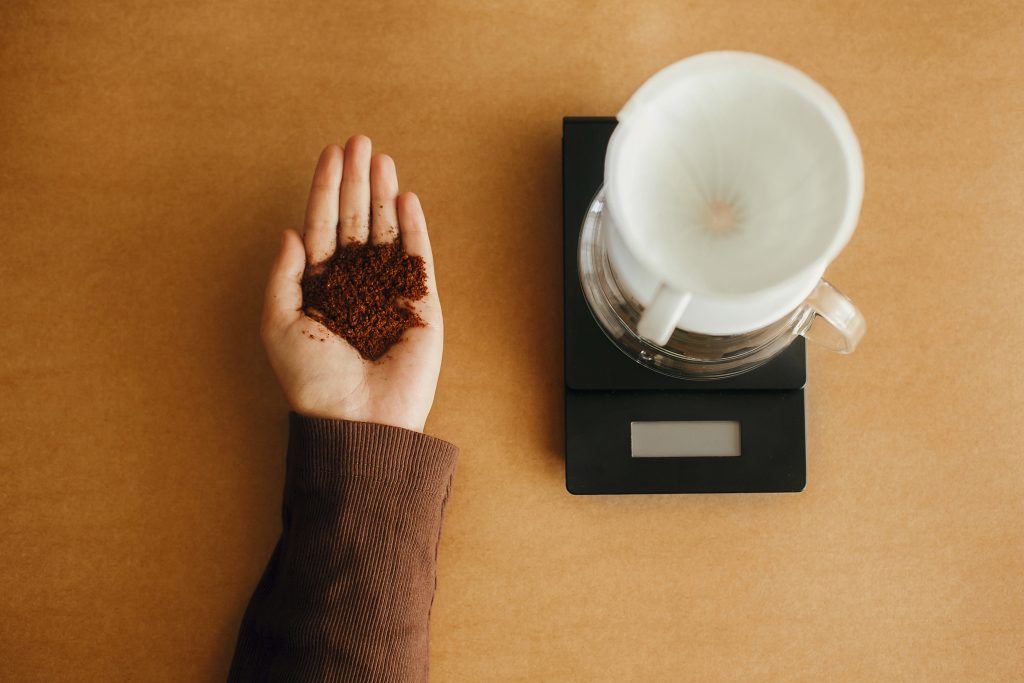 Grind coffee close up in hands on background of glass kettle with pour over on scale, flat lay. Preparing for alternative coffee brewing. Hand holding grounded coffee on brown background.